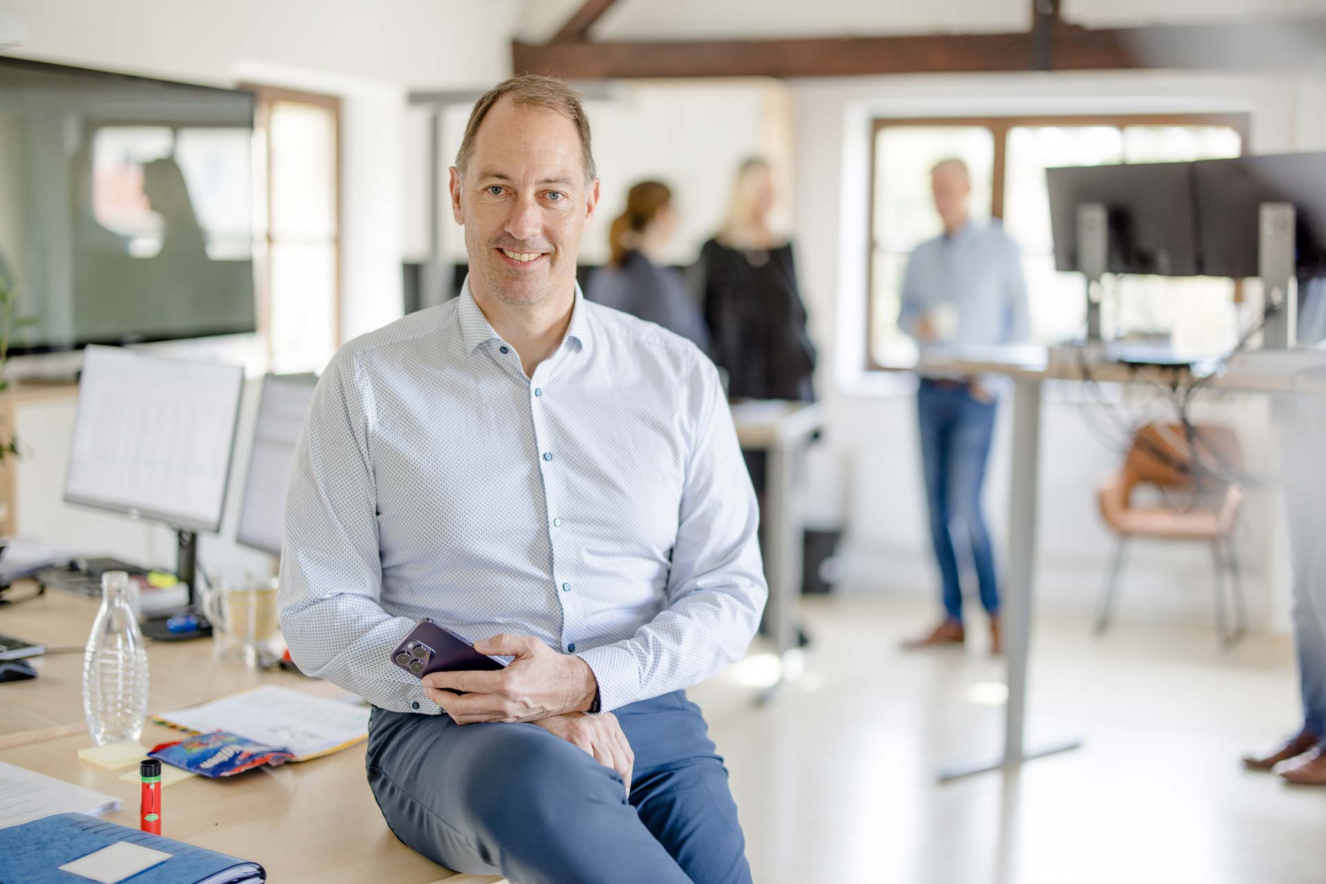 Geschäftsführer Klaus Busch sitzt auf einem Tisch und hält ein Handy in der Hand. Im Hintergrund sieht man in der Unschärfe das Büro und weitere Kollegen